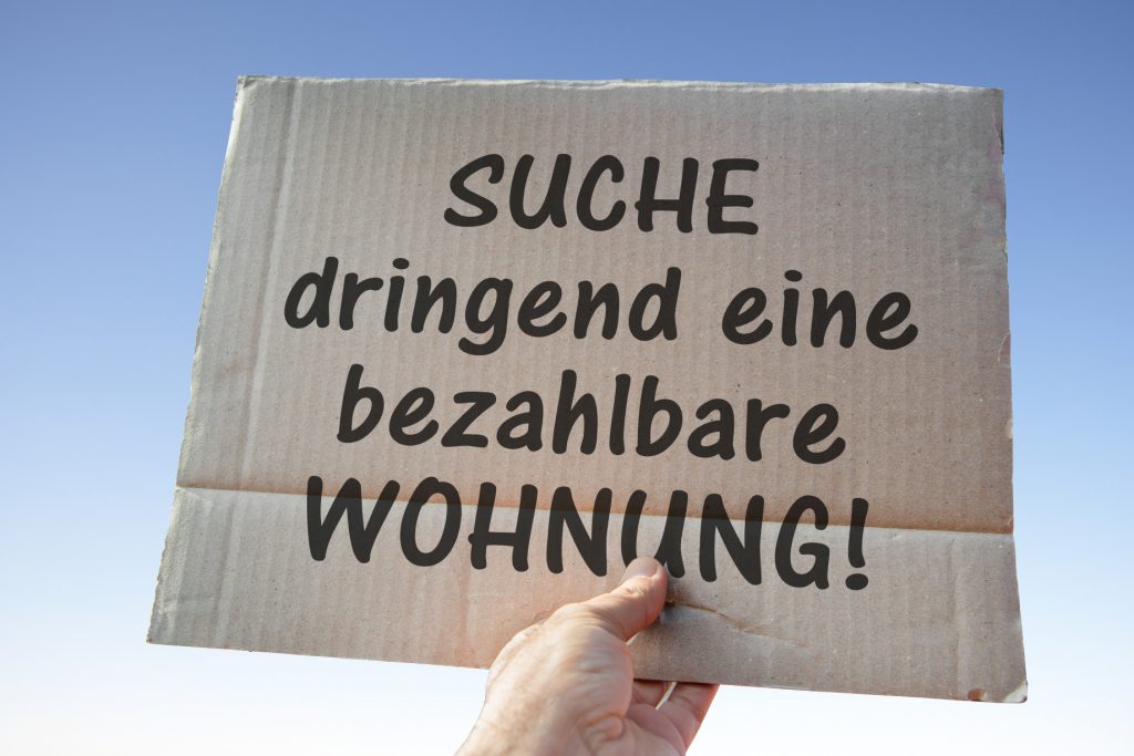 Hand hält Pappschild mit der Aufschrift „Suche dringend eine bezahlbare Wohnung“ vor blauem Himmel