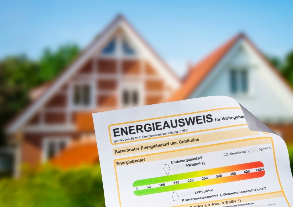 Energieausweis mit farbiger Effizienzskala vor einem Wohnhaus im Hintergrund
